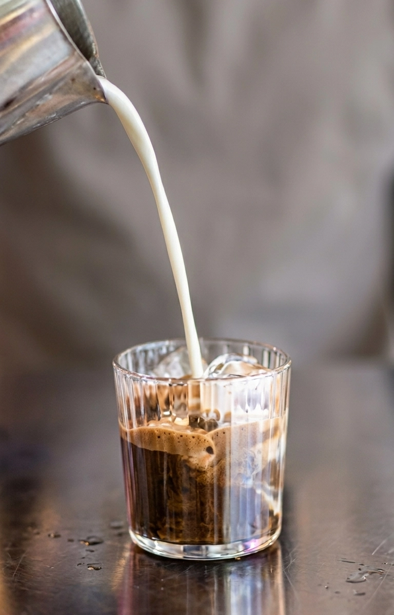 Milch wird aus einer Metallkanne in ein geripptes Glas mit Hojicha-Espresso gegossen – Zubereitung eines Hojicha Latte auf einer Metalltheke