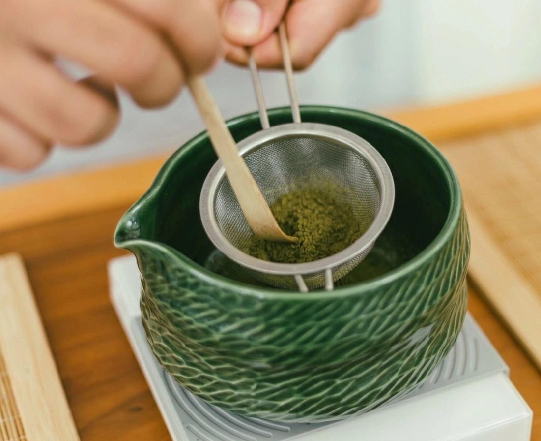 Hojicha-Pulver wird durch ein feines Metallsieb mit einem Bambuslöffel in eine grüne Keramik-Chawan gesiebt – traditionelle Teezubereitung auf einem Holztablett