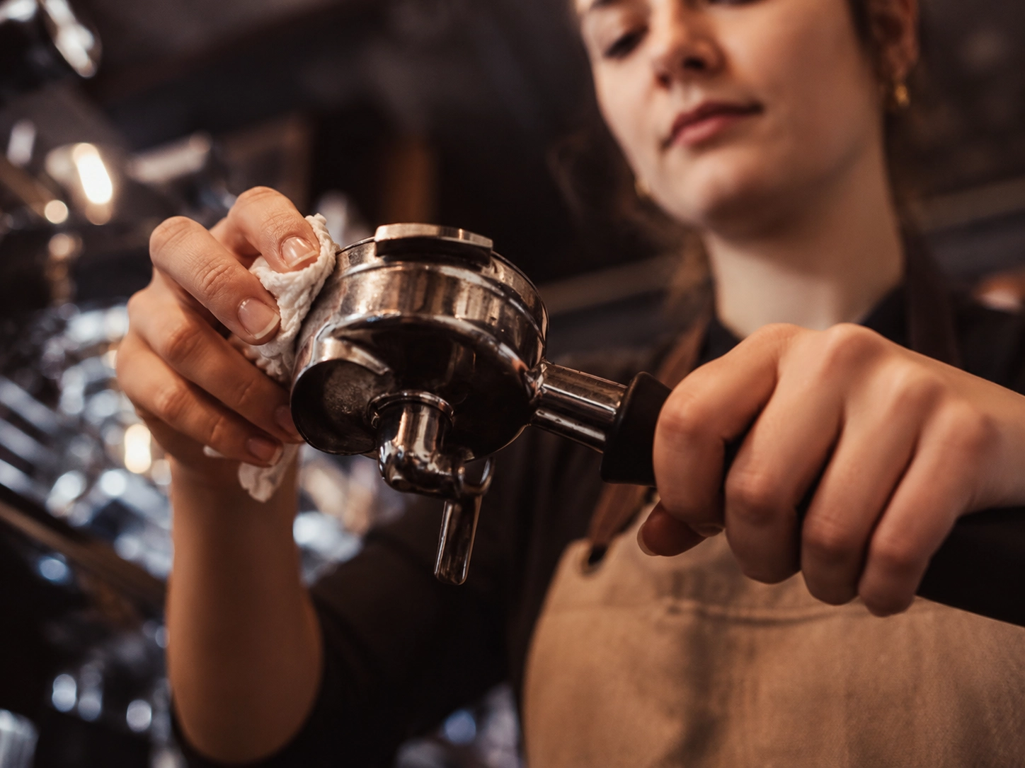 Nahaufnahme einer Barista-Frau, die den Siebträger einer Espressomaschine sorgfältig mit einem Tuch reinigt.
