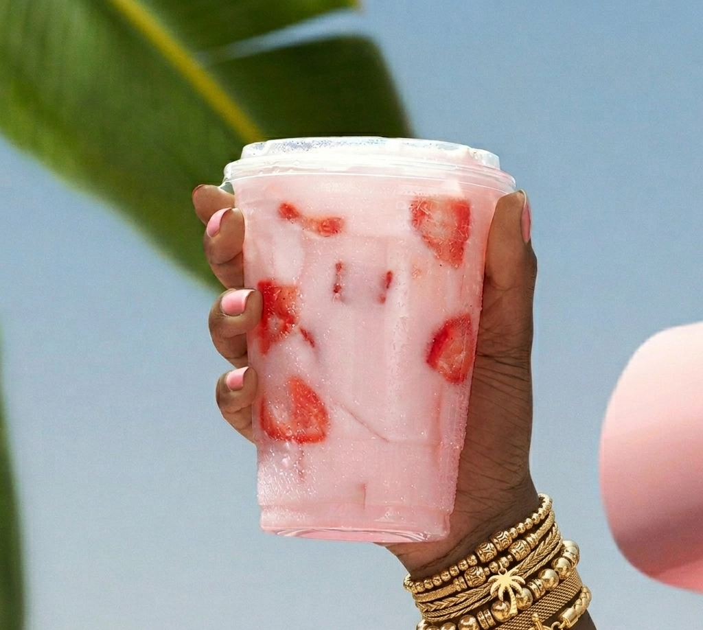 Pink Drink im To-go-Becher mit frischen Erdbeerstücken, Crushed Eis und cremiger Kokosmilch, gehalten gegen blauen Sommerhimmel
